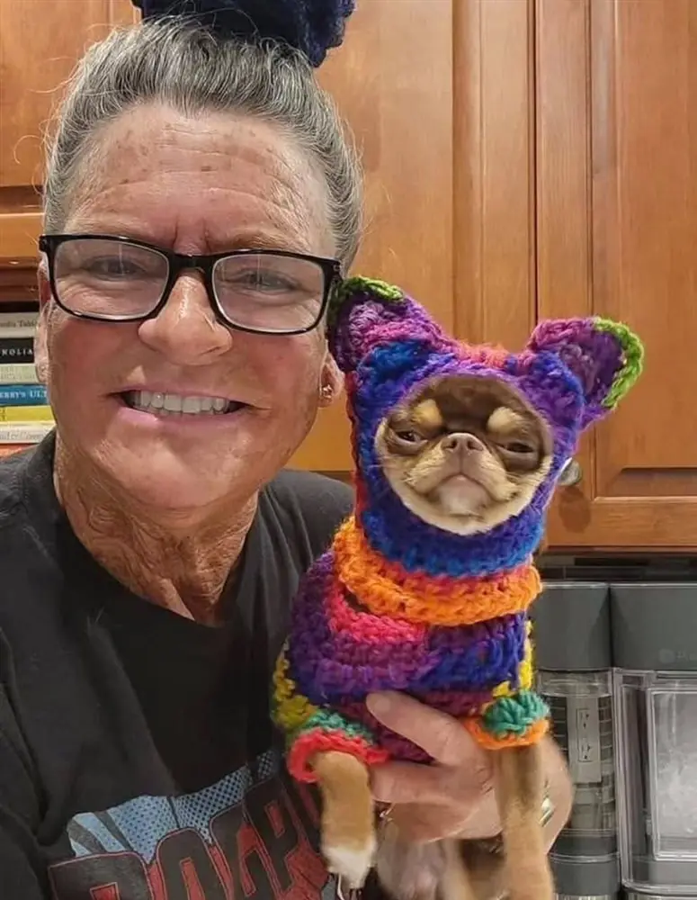 The Diamond Chihuahuas breeder smiling and holding an adult Chihuahua in a rainbow knitted sweater and hat in her kitchen
